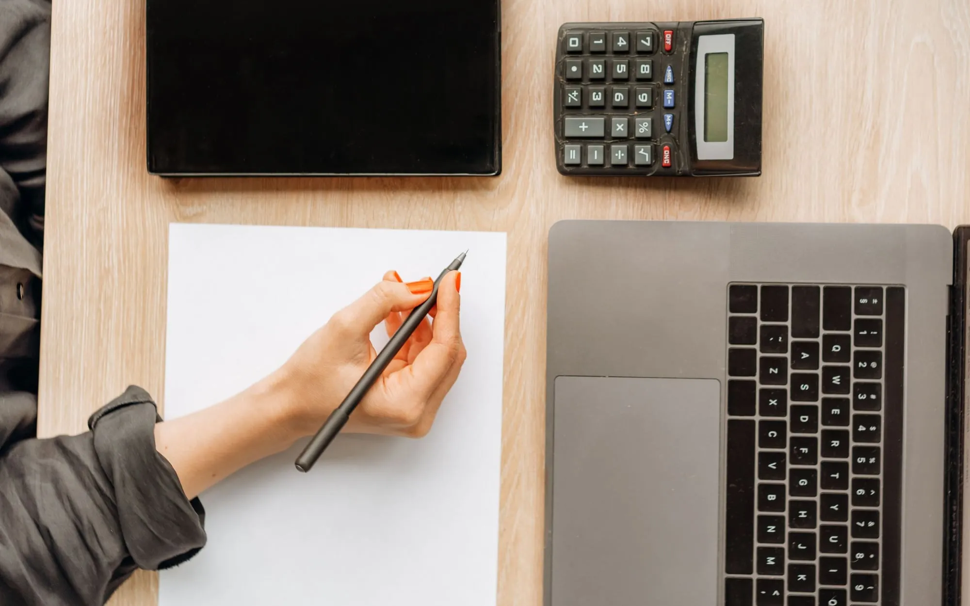 A person writing notes beside a laptop, a tablet and a calculator on a desk