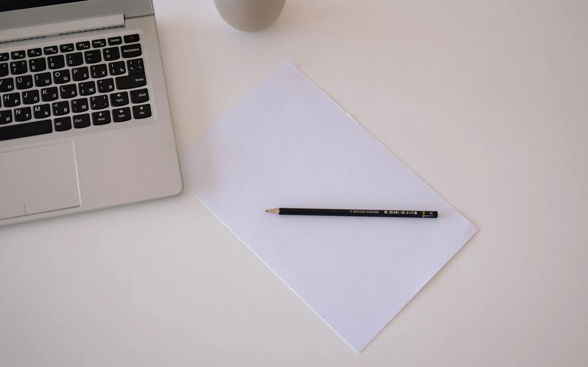 A laptop, a sheet of paper and a pencil on a clean white desk, seen from above